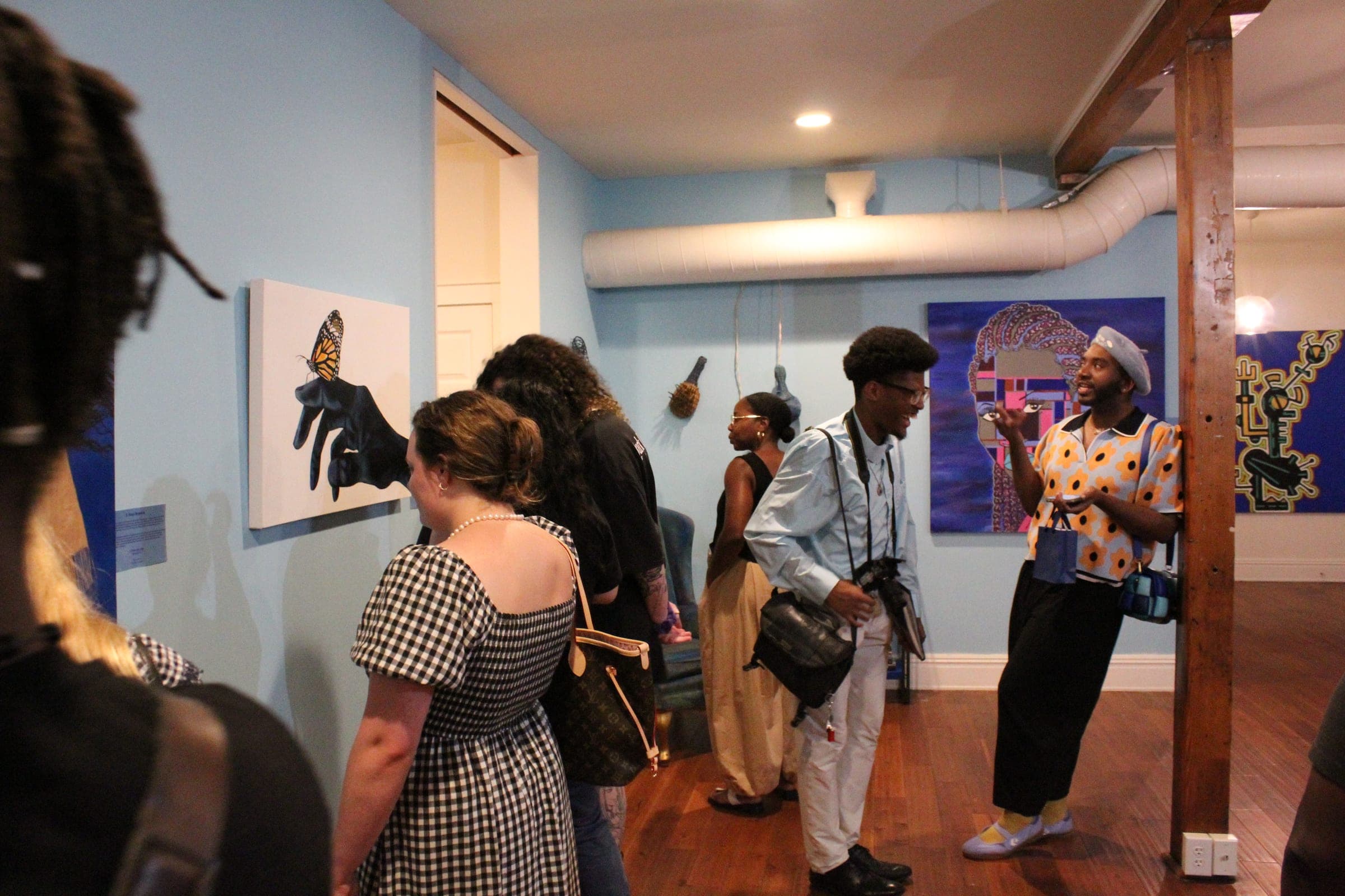 Visitors fill a blue-walled gallery room hung with paintings, including one of a butterfly resting on a gloved hand