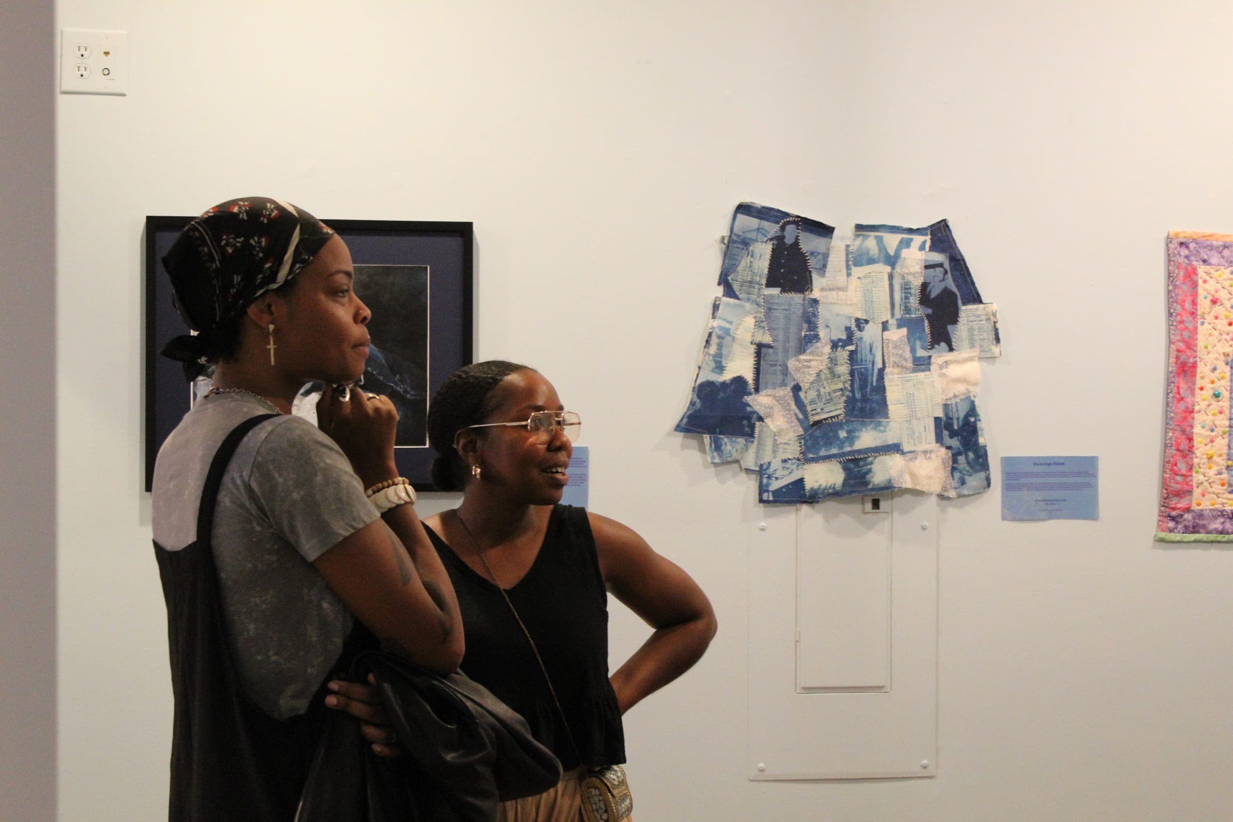 Two women study a cyanotype collage of photographs and fabric hung on a white gallery wall