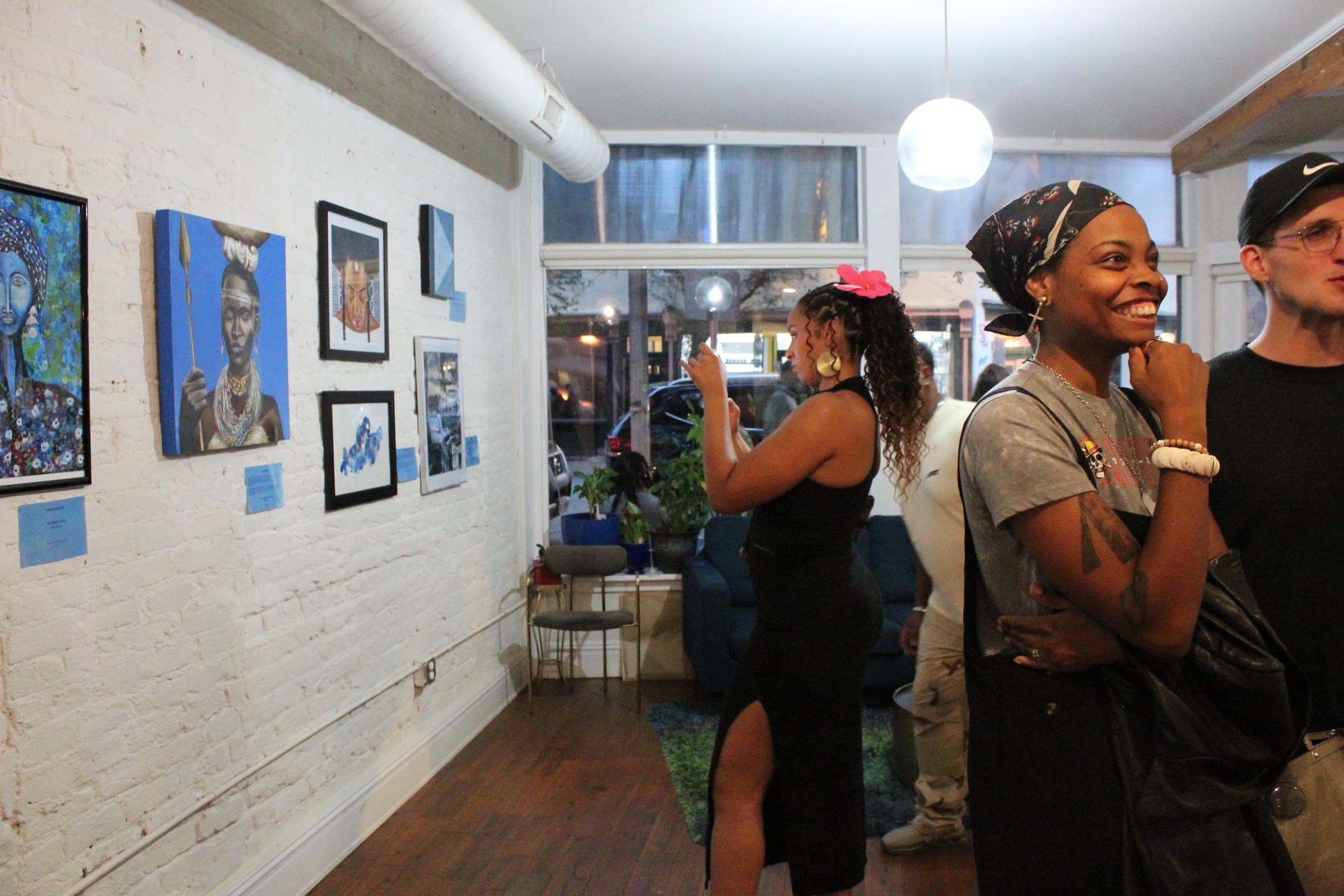 Visitors talk and take photos along a white-brick wall hung with paintings, with the street visible through the front windows