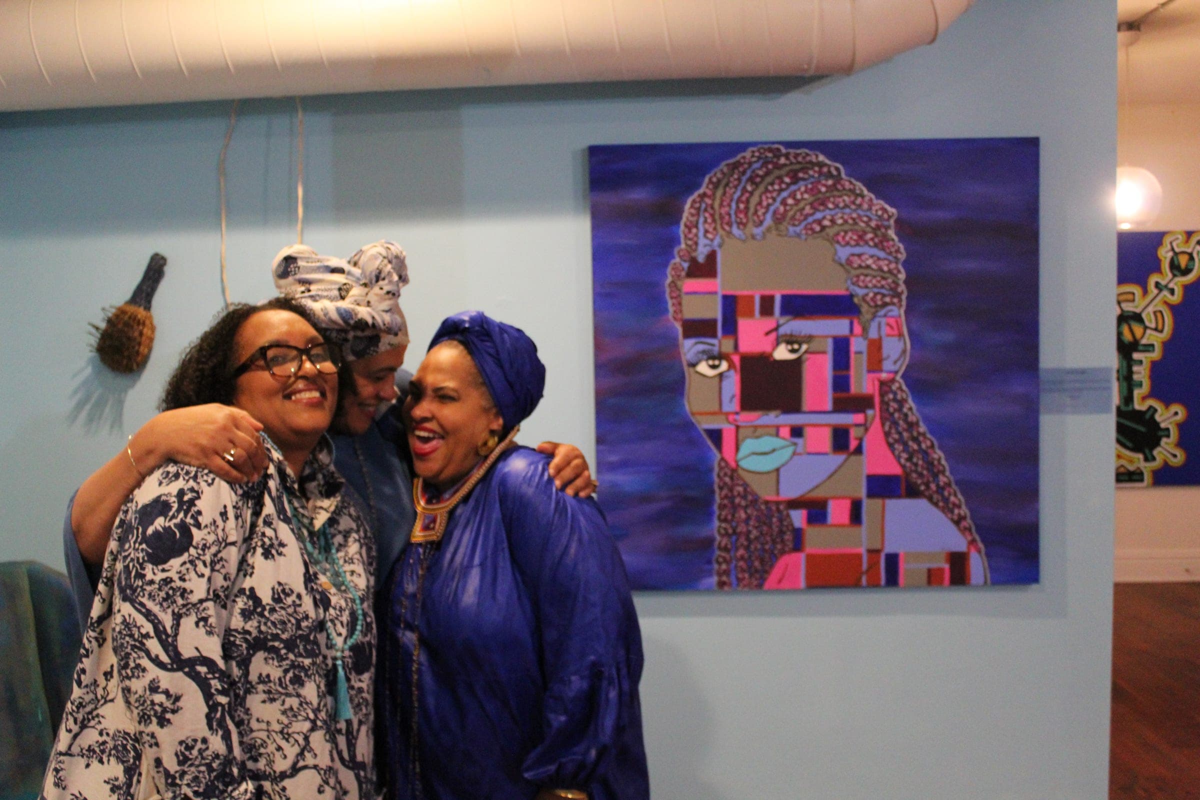 Three women embrace and laugh in front of a large portrait painted in pink, violet, and blue