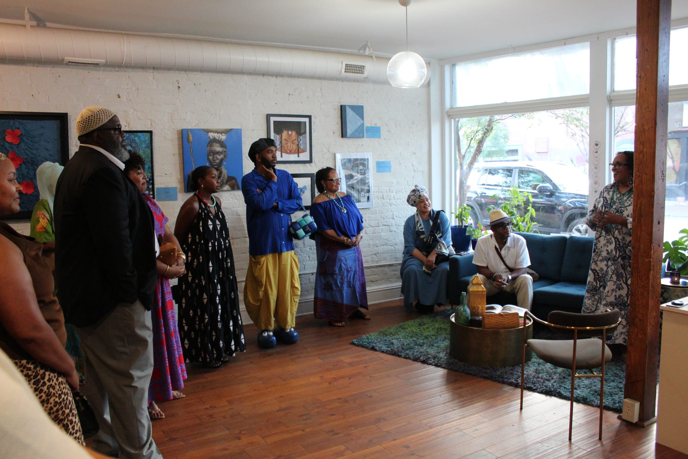 Guests gather in a loose circle in a white-brick gallery hung with paintings, beside a blue sofa and tall front windows