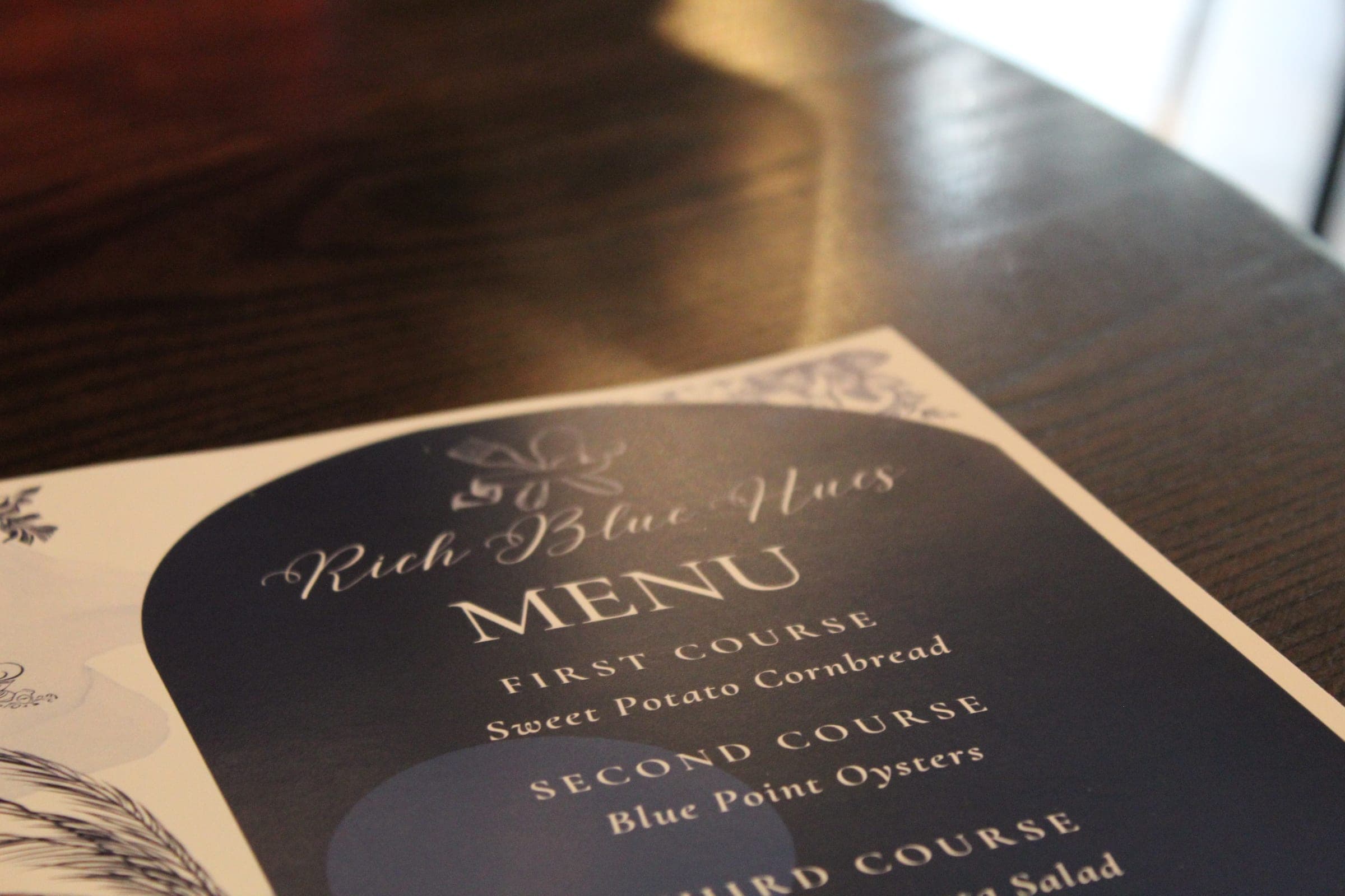 A printed Rich Blue Hues menu listing sweet potato cornbread and Blue Point oysters, on a dark wood table