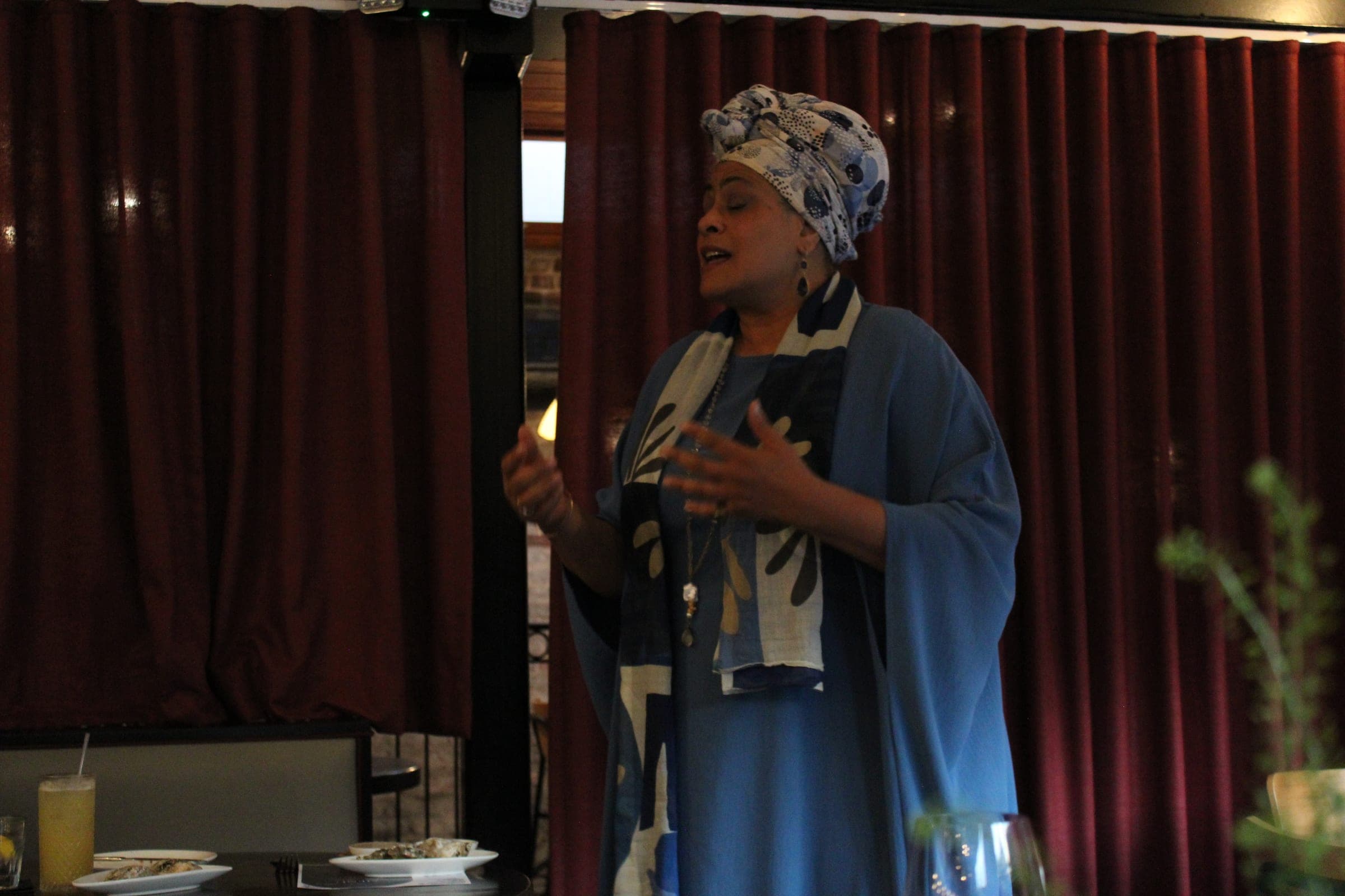 A woman in a blue head wrap and flowing blue robe speaks to diners in front of a deep red curtain